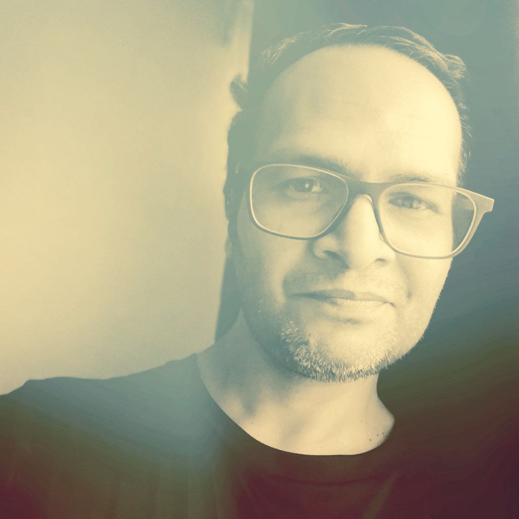 A close-up portrait of a man with glasses, smiling gently, featuring a soft, vintage-style filter.